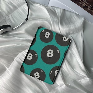 May include: A rectangular teal-colored item featuring a pattern of black billiard balls with the number 8 in white. The item is resting on a white, silky surface, with a pair of headphones visible in the background.