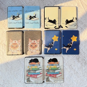 May include: Five e-reader cases featuring various cat illustrations. One shows a black cat on a book with the text 'Life is better with Cats and books.' Others depict a black cat on a desk, a cat in a strawberry hood, a cat reaching for a star, and a cat on a stack of books.