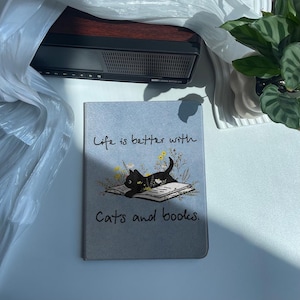 Op de afbeelding: Een grijze en blauwe notitieboekomslag met een zwarte kat die een boek leest. De omslag heeft de tekst "Life is better with cats and books."