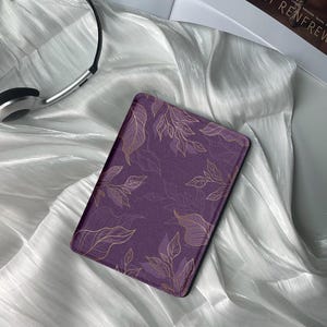 May include: A purple e-reader case with a gold floral design. The case is lying on a white silky fabric.