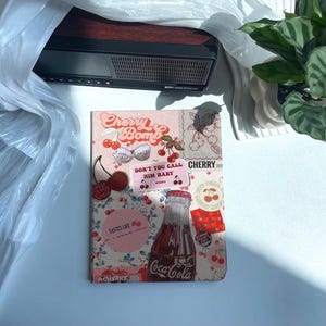 Könnte beinhalten: Ein rosa-weißes Notizbuch mit einer Collage aus Kirschen, einer Coca-Cola-Flasche und dem Text "Cherry Bomb" und "Don't You Call Him Baby".