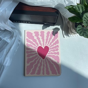 May include: A pink and white journal with a heart design. The journal features a large pink heart in the center, surrounded by radiating lines. The cover has a textured appearance, and the overall design is simple and eye-catching.