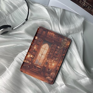 May include: A brown leather e-reader case with a whimsical illustration of a library with a large window and glowing lights. The case is lying on a white fabric surface.