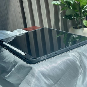 May include: A black iPad with a white stylus on a gray fabric surface. The iPad is closed and the stylus is lying on top of it.