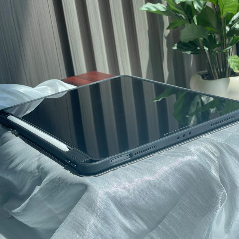 May include: A black iPad with a white stylus on a gray fabric surface. The iPad is closed and the stylus is lying on top of it.