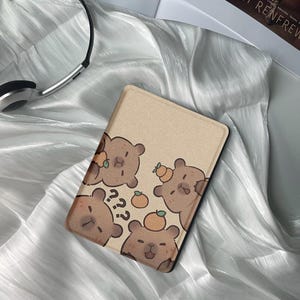 May include: A tan tablet case featuring cartoon brown bears with oranges. The bears have question marks and are in various poses. The case has a black border and is on a white, silky surface.