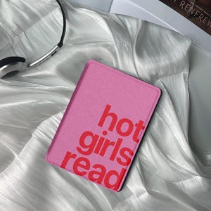 May include: A pink e-reader cover with the words "hot girls read" in red. The cover is rectangular and has a black border. The e-reader cover is on a white, shiny fabric. Silver headphones are in the background.