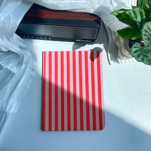 Può includere: Un taccuino rettangolare con un motivo a righe rosse e rosa. Il taccuino è appoggiato su una superficie bianca. Sullo sfondo c'è un altoparlante nero e marrone, parzialmente oscurato da un sacchetto di plastica. È visibile anche una pianta in vaso.