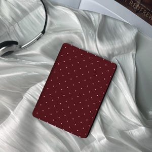 Puede incluir: Una funda de tableta burdeos con un patrón de lunares blancos. La funda rectangular tiene esquinas redondeadas. Se ven unos auriculares plateados en el fondo.