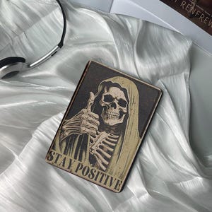 Peut inclure: Une image en noir et blanc d'un squelette portant une robe à capuche donnant un pouce en l'air. Le texte "STAY POSITIVE" est imprimé en bas de l'image.