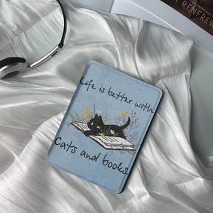 Könnte beinhalten: Ein blauer und weißer Buchumschlag mit einer schwarzen Katze, die ein Buch liest. Der Text auf dem Umschlag lautet "Life is better with cats and books."