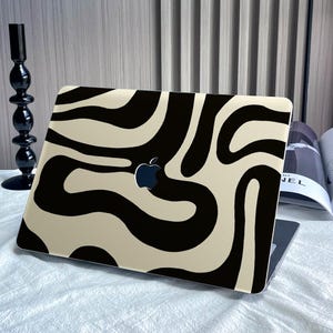 Puede incluir: Un ordenador portátil con una piel de patrón abstracto negro y beige. El patrón se asemeja a una impresión de cebra con líneas onduladas.