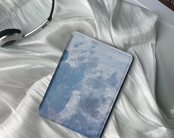 May include: A rectangular e-reader device featuring a protective cover with an abstract blue and white painted texture. The device is positioned alongside a pair of black and silver over-ear headphones, showcasing a sleek and functional design.