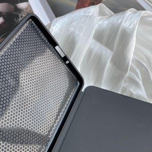 May include: A black tablet case with a honeycomb pattern on the inside cover. The case is open, revealing the interior design. The background features a white, flowing fabric.