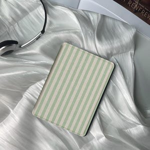 以下が含まれることがあります： ミントグリーンと白のストライプ模様の長方形の電子書籍リーダーケース。ケースは白くシルキーな生地の上に置かれています。シルバーのヘッドフォンが左上にあります。