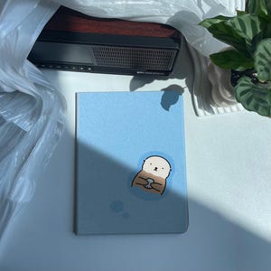 Könnte beinhalten: Ein hellblaues Notizbuch mit einem Cartoon-Seeotter, der eine Muschel hält. Der Otter ist von Blasen umgeben. Das Notizbuch liegt auf einer weißen Oberfläche.