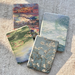 May include: Four rectangular e-reader cases featuring printed fine art reproductions. One case displays a sunset landscape, another a water lily pond, a third a different water lily scene, and the fourth depicts white almond blossoms on blue.
