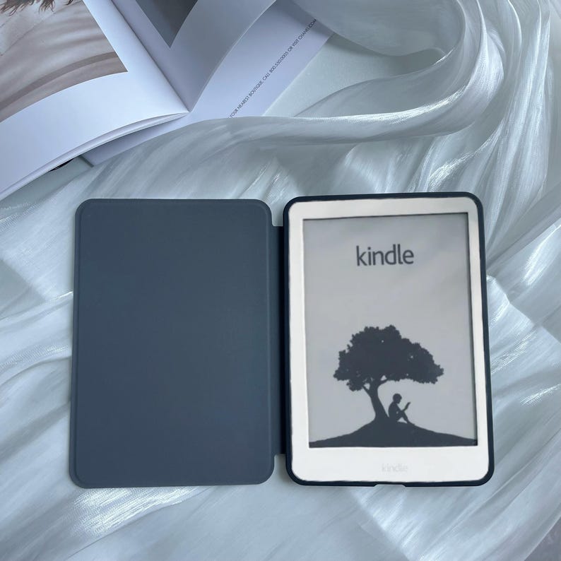 May include: A gray Kindle e-reader with a black cover and a white screen displaying the word "kindle" and a silhouette of a tree with a person sitting under it.