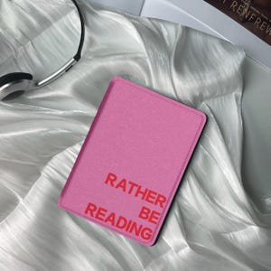 Peut inclure: Une housse rose pour liseuse avec le texte "Rather Be Reading" en rouge.