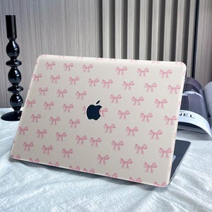 Pode incluir: Uma pele para laptop de cor creme com um padrão repetitivo de laços cor-de-rosa. O logotipo da Apple é visível na pele.