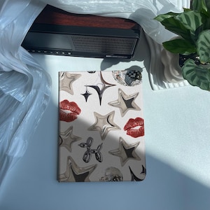 Puede incluir: Un cuaderno decorativo con un diseño de estrellas plateadas, labios rojos y un perro globo sobre un fondo blanco. Una bola de discoteca y otras formas de estrella completan el diseño. El cuaderno está sobre una superficie blanca.