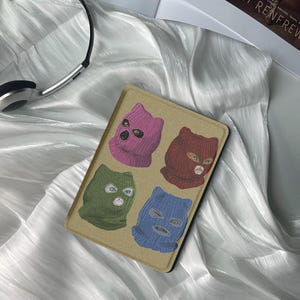 May include: A brown and beige patterned cover for an e-reader with four cats wearing different colored ski masks. The cats are wearing pink, red, green, and blue ski masks.