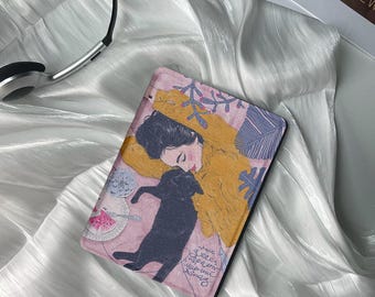 May include: A pink and blue illustrated case for an e-reader featuring a woman with black hair and a black cat. The woman is wearing a yellow sweater and the text "make every moment magical" is written on the case.