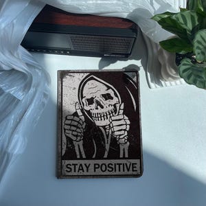 Könnte beinhalten: Ein rechteckiges Metallschild mit abgenutztem Finish zeigt einen Schädel mit Kapuze, der den Daumen hochhält. Die Worte "STAY POSITIVE" sind unten in fetter Schrift gedruckt. Das Schild hat eine Vintage- und Gothic-Ästhetik.