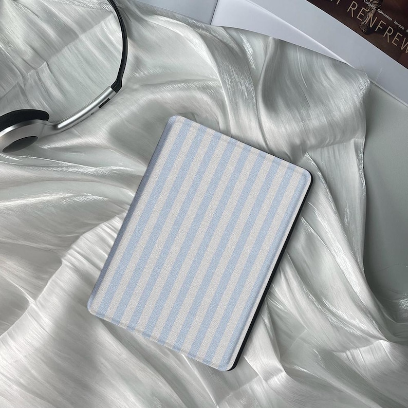 May include: A rectangular e-reader case with a light blue and white striped pattern. The case is resting on a shiny, white, silky surface. A pair of silver headphones is visible in the upper left corner.