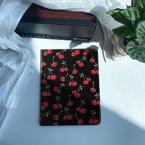 May include: A black and red cherry print notebook cover. The cover is made of a durable material and features a repeating pattern of cherries.