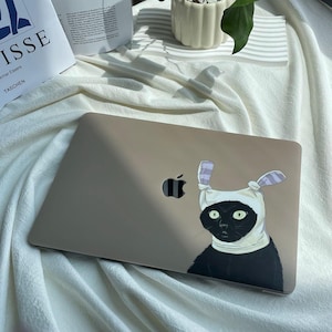 Pode incluir: Um laptop prateado com um adesivo de um gato preto vestindo uma fantasia de coelho branco na tampa. O gato tem olhos verdes e orelhas pretas. O laptop está em uma superfície de tecido branco.