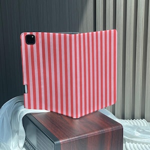 Può includere: Una custodia per tablet con un motivo a righe verticali in tonalità alternate di rosso e rosa chiaro. La custodia presenta un ritaglio per l'obiettivo della fotocamera nell'angolo in alto a sinistra e una fessura laterale per riporre un pennino.