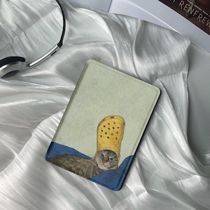 May include: A tablet case featuring a cat wearing a yellow clog shoe as a hat. The cat is gray and brown, resting on a blue surface, with a light green background. The case has a black border.