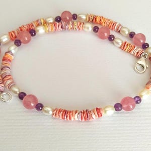 May include: A necklace featuring a combination of pink, white, and purple beads. The necklace includes pink gemstones, white pearls, and small, colorful shell-like beads. It has a silver clasp.