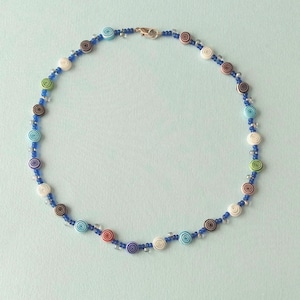 May include: A beaded necklace with a blue cord and a variety of colorful spiral-patterned beads. The beads are in shades of white, brown, blue, green, and red. The necklace is secured with a silver clasp.