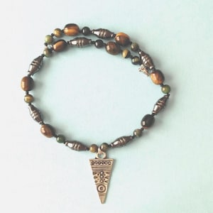 May include: A handcrafted necklace featuring a mix of brown, green, and metallic beads. The necklace has a pendant with an etched design. The beads are various shapes and sizes, creating a unique and textured look.