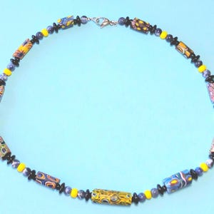 May include: A necklace with a variety of colorful beads. The necklace features cylindrical beads with intricate patterns in yellow, blue, and red, interspersed with smaller black, yellow, and blue beads. The necklace is on a light blue background.