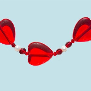 May include: A red heart-shaped bead necklace with white pearl accents. The necklace is made of glass beads and is strung on a clear thread.