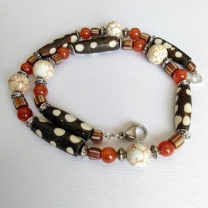 May include: A beaded necklace featuring dark brown cylindrical beads with white polka dots, round white marbled beads, translucent orange spherical beads, and small striped cylindrical beads, all separated by silver-toned metal spacers with a lobster clasp.