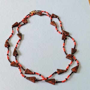 May include: A necklace featuring triangular brown pendants interspersed with red and black beads. The necklace has a gold-colored clasp and is displayed against a light blue background. The beads are small and evenly spaced.