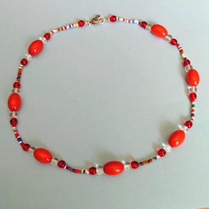 May include: A necklace featuring large, oval red beads interspersed with smaller clear and red beads. The necklace also includes a strand of tiny, colorful beads, and a silver clasp. The jewelry is displayed on a light blue background.