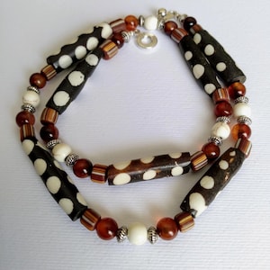 May include: A two-strand necklace featuring a mix of beads. The necklace includes dark brown cylindrical beads with white polka dots, interspersed with white, brown, and reddish-brown beads. The necklace is secured with a silver-colored clasp.