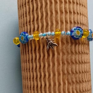 May include: A beaded bracelet featuring blue and yellow floral beads, interspersed with yellow and clear beads. A silver-colored bee charm dangles from the bracelet. The bracelet is displayed against a textured, brown background.