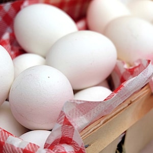 May include: A basket filled with white eggs on a red and white checkered cloth. The eggs are arranged in a single layer, with some overlapping.