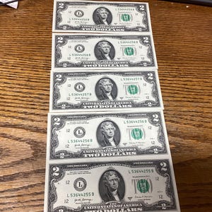 May include: Five United States two-dollar bills are arranged on a wooden surface. Each bill features a portrait of Thomas Jefferson and the words "Two Dollars". The bills are green and black.