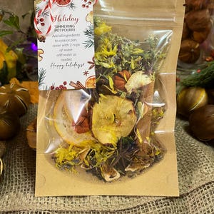 May include: A clear cellophane bag filled with dried potpourri, including orange slices, apple pieces, and star anise. A tag with the words "Holiday Simmering Potpourri" is attached with a red and white string. The tag also includes instructions and the phrase "Happy Holidays!"