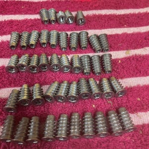 May include: A collection of silver-colored, threaded furniture inserts arranged on a pink and white striped surface. The inserts have a flanged head and a ribbed body. A string of white lights with a red bulb is in the background.