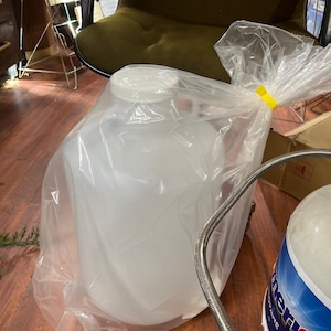 May include: A large, white, plastic container with a screw-on lid, sealed inside a clear plastic bag. The bag is secured with a yellow twist tie. A metal hose and a portion of a blue and white tank are visible.