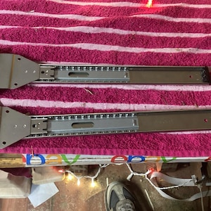 May include: Two silver metal drawer slides with mounting brackets. The slides are designed for smooth drawer operation and are made of durable metal. They are shown on a pink and white striped towel.