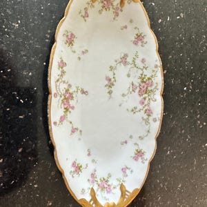 May include: An oval porcelain serving dish featuring a white base decorated with small pink floral clusters and green vines. The scalloped edges are trimmed with gold, and the two ends feature ornate, raised gold-painted decorative accents.